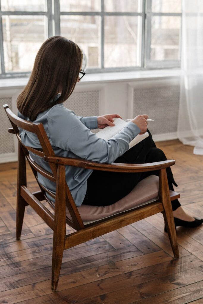 Woman with glasses sitting in a wooden chair while writing in a notebook, capturing thoughtful moments.